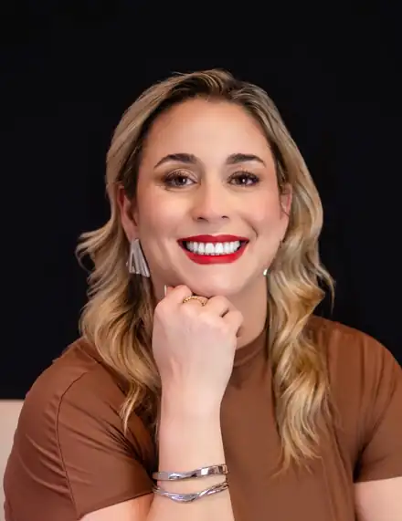 Carol Modesto sorrindo, com batom vermelho, apoiando o queixo na mão